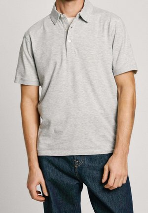 Mann trägt hellgraues kurzärmeliges Poloshirt und dunkelblaue Jeans, steht mit locker hängenden Armen seitlich vor einfarbigem Hintergrund.