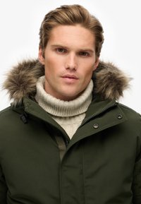 Parka vert avec une capuche bordée de fausse fourrure, dotée d'une fermeture éclair complète et d'une fermeture à pression. Superposée sur un col roulé côtelé beige clair.