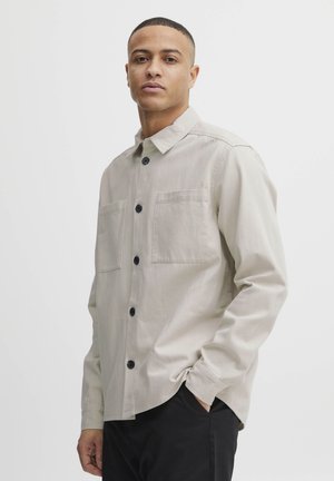 Light beige button-up shirt with two front pockets, black buttons, and a classic collar. The fabric appears smooth and slightly textured.