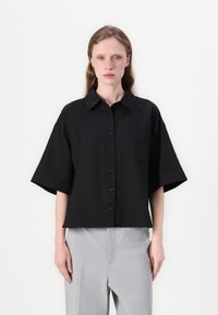 Camisa preta de manga curta em um tecido leve. Apresenta fecho de botão na frente, colarinho e um único bolso no peito. Combinada com calças cinzentas claras.