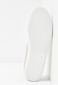 Baskets blanches avec une semelle texturée, présentant un dessus tissé en beige et des accents métalliques, inclinées contre un fond blanc uni.