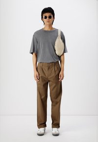 Gray oversized t-shirt, brown pleated trousers, beige sling bag, and white sneakers with black accents. Smooth fabric, casual fit, neutral colors.