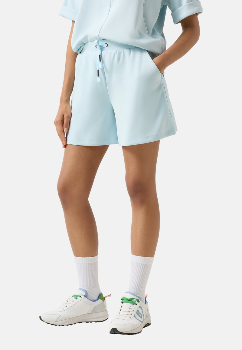 Personne portant un short bleu clair à cordon avec poches, des chaussettes blanches mi-mollet et des baskets blanches à lacets verts sur un fond uni.