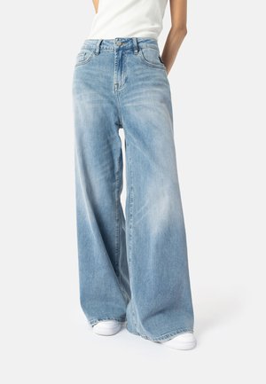 Lyseblå vid bukse laget av denim, med høy midje, fem lommer, og en lett falmet tekstur. Parret med hvite joggesko.