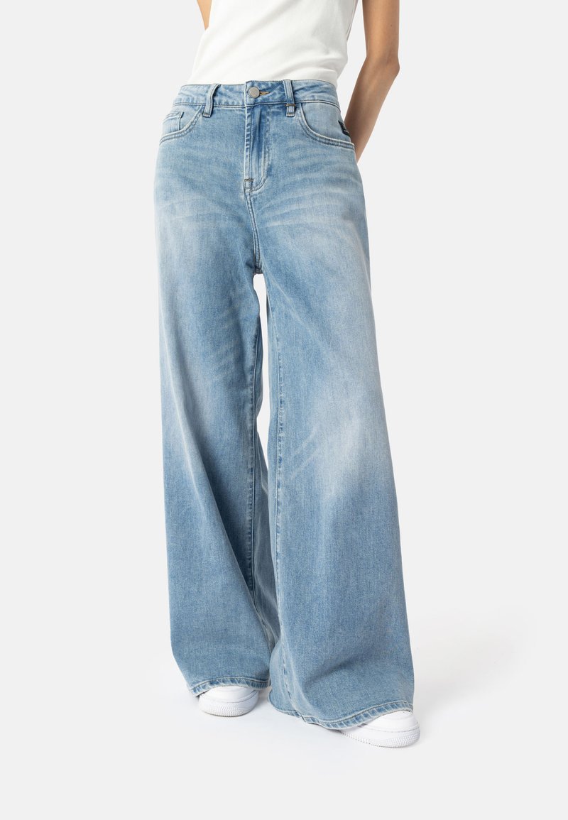 Svetlomodré široké džínsy vyrobené z denimu, s vysokým pásom, piatimi vreckami a mierne vybledlou textúrou. Skombinované s bielymi teniskami.