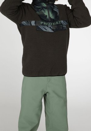 Fleece pullover i mørkebrun med et blankt sort brystpanel og grønt logo, parret med lysegrønne bukser, der har en afslappet pasform.