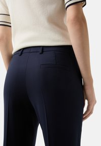 Pantalons taillés marine avec un devant plat, des poches latérales et une poche arrière passepoilée unique. Le matériau semble lisse avec un léger brillant.