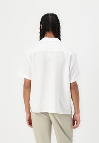 Camicia bianca a maniche corte con un motivo testurizzato, colletto classico e vestibilità rilassata, indossata con pantaloni chiari.