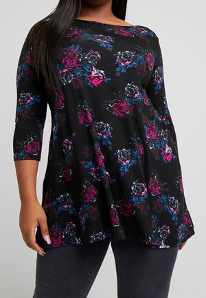 Woman wearing a black tunic with pink, purple, and blue floral patterns and three-quarter sleeves, paired with dark pants.