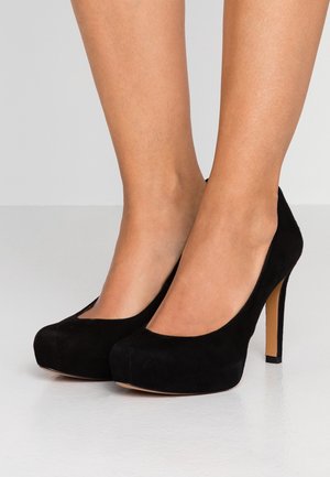 Füße in schwarzen Wildleder-Pumps mit hohen Absätzen, die auf einer glatten, hellen Oberfläche stehen, vor einem schlichten Hintergrund.