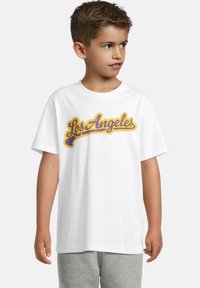Ung gutt med kort hår iført en hvit T-skjorte med "Los Angeles" i lilla og gul skrift, kombinert med grå bukser, ser mot høyre.