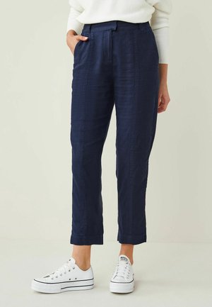 Kobieta ubrana w granatowe spodnie do kostek z przodu z plisami i mankietami, w połączeniu z białymi niskimi trampkami i białym swetrem.