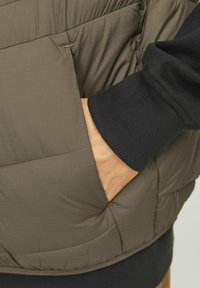 Olivgrüner Puffer-Weste mit diagonaler Naht, strukturiertem Stoff und Seitentaschen. Schwarze gerippte Ärmelabschlüsse sind sichtbar.