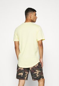 Gelbes T-Shirt mit kurzen Ärmeln und abgerundetem Saum, kombiniert mit Shorts im Tarnmuster in verschiedenen Grüntönen und Braun.