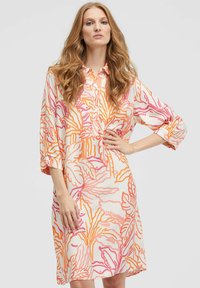 Frau mit langen, gewellten Haaren, die ein tailliertes knielanges Kleid mit orange- und pinkfarbenen abstrakten Blumenmustern trägt und mit einer Hand auf der Hüfte posiert.