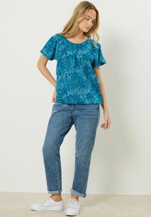 Nainen pukeutuneena siniseen kuvioituun lyhythihapaidan, käärityt siniset farkut ja valkoiset tennarit, seisomassa yksinkertaista vaaleaa taustaa vasten.