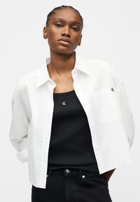 Νεαρή γυναίκα που φοράει ένα λευκό πουκάμισο με κουμπιά πάνω από ένα μαύρο τοπ και μαύρο παντελόνι, και τα δύο με μικρά λογότυπα "cK".