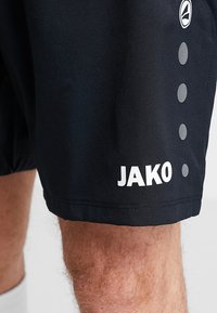 Schwarze Sportshorts aus glattem Stoff mit einem weißen "JAKO"-Logo und runden Musterdetails entlang der Seitennaht.