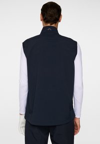 Marinblå ärmlös väst med hög krage, tillverkad av ett slätt tyg, med en logotyp på baksidan och en avslappnad passform för rörelsefrihet.