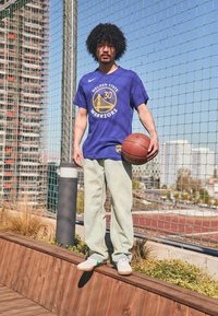 Tricou scurt cu mânecă albastră, purtând logo-ul "Golden State Warriors", pantaloni verzi deschis, ținând o minge de baschet, stând pe un zăgaz din lemn.