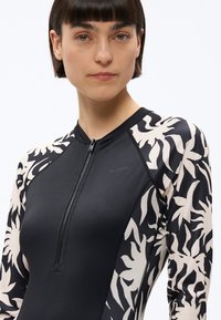 Žena s krátkymi hnedými vlasmi, oblečená v dlhom rukáve plaviek s čierno-krémovým kvetinovým vzorom a predným zipsom, stojaca pred bielym pozadím.