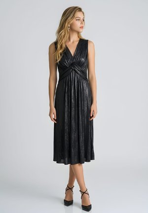 Joven vestida con un vestido midi negro brillante sin mangas con pliegues y detalle cruzado en la cintura, combinado con tacones negros de tiras, mirando hacia la derecha.