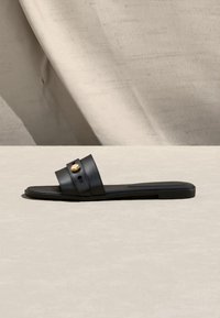 Μαύρο επίπεδο σανδάλι σλάιντ με φαρδιά λουρί που διαθέτει ένα χρυσό κυκλικό κουμπί, παρουσιασμένο σε ανοιχτό μπεζ επιφάνεια και φόντο.