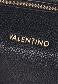 Crna kožna torbica s teksturom koja sadrži zlatno slovo "VALENTINO" i zlatni patentni zatvarač. Površina ima uzorak kamenčića.