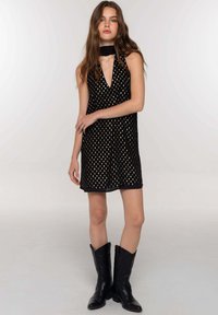 Giovane donna che indossa un vestito nero senza maniche con un motivo dorato, una sciarpa nera e stivali da cowboy neri, in piedi contro uno sfondo bianco.