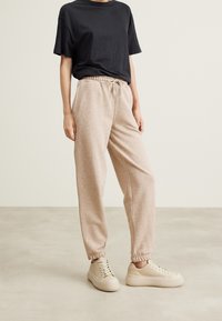 Beżowe spodnie dresowe z elastycznymi ściągaczami i talią na sznurek, połączone z krótkim czarnym t-shirtem i beżowymi trampkami na neutralnym tle.