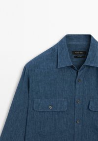 Camicia blu a maniche lunghe con bottoni, dotata di due tasche frontali con pattina e bottone, colletto a punte e etichetta visibile all'interno del colletto.