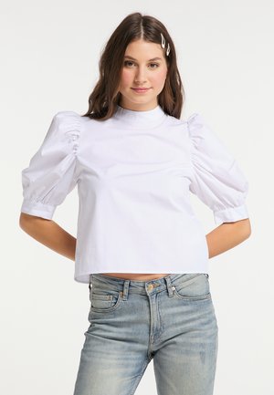 Giovane donna con lunghi capelli castani che indossa una camicetta bianca con maniche a sbuffo e jeans azzurri, in piedi con le mani dietro la schiena.
