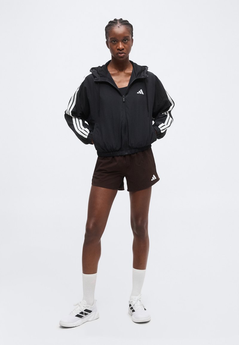 Giovane donna in piedi con le mani nelle tasche, indossa una giacca nera Adidas, pantaloncini scuri, calzini bianchi e scarpe da ginnastica bianche.