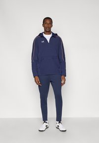 Μπλε σκούρο πουλόβερ με φερμουάρ μέχρι τη μέση, πορτοκαλί λεπτομέρειες και μπροστινή τσέπη. Σετάρεται με αντίστοιχο μπλε σκούρο παντελόνι και λευκά αθλητικά παπούτσια με μαύρες λεπτομέρειες.