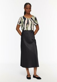 Kurzärmeliges Oberteil mit einem abstrakten Muster in Grün, Pink und Schwarz; kombiniert mit einem langen, schwarzen Satinskirt und schwarzen Ballerinas.