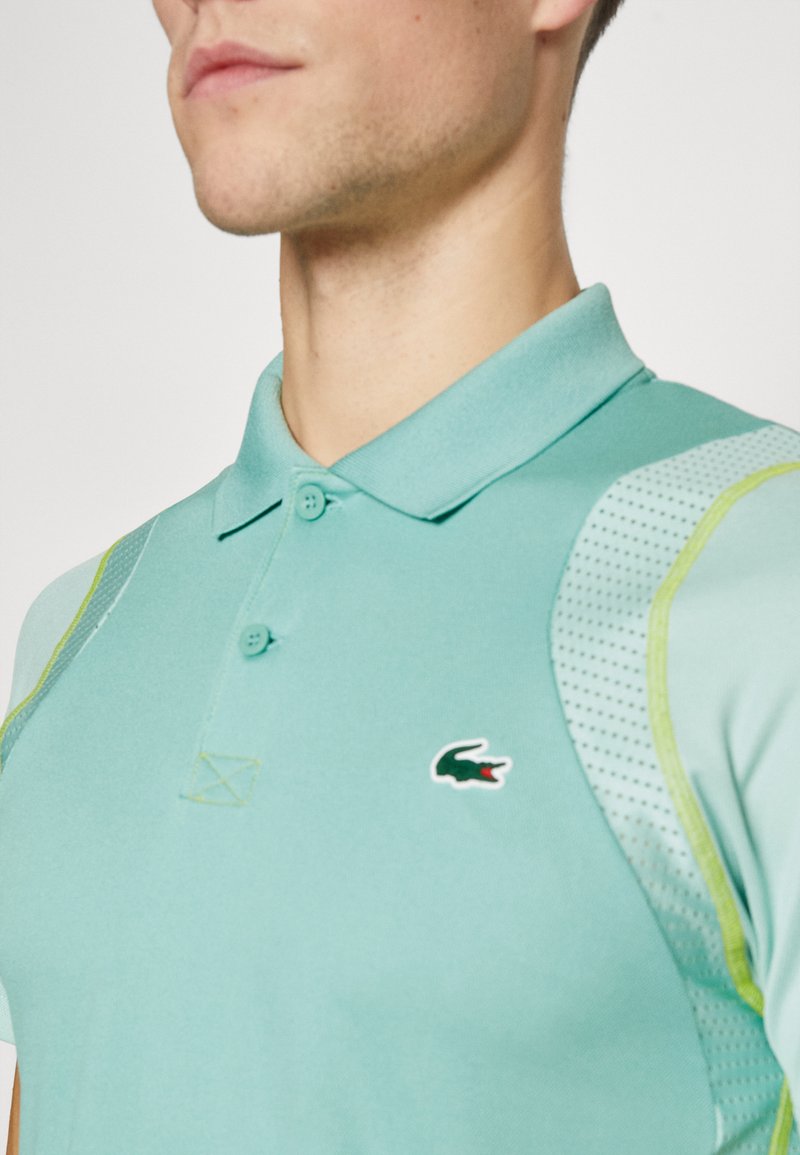 Polo bleu clair avec un col classique, présentant une accent vert sur l'épaule et un petit logo de crocodile sur la poitrine.