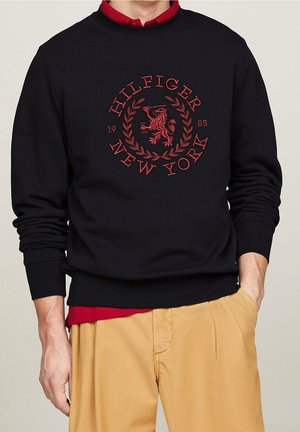 Muž v černé mikině s červeným emblémem „Hilfiger New York 1985“, s červenou košilí pod ní a béžovými kalhotami, jednou rukou v kapse.
