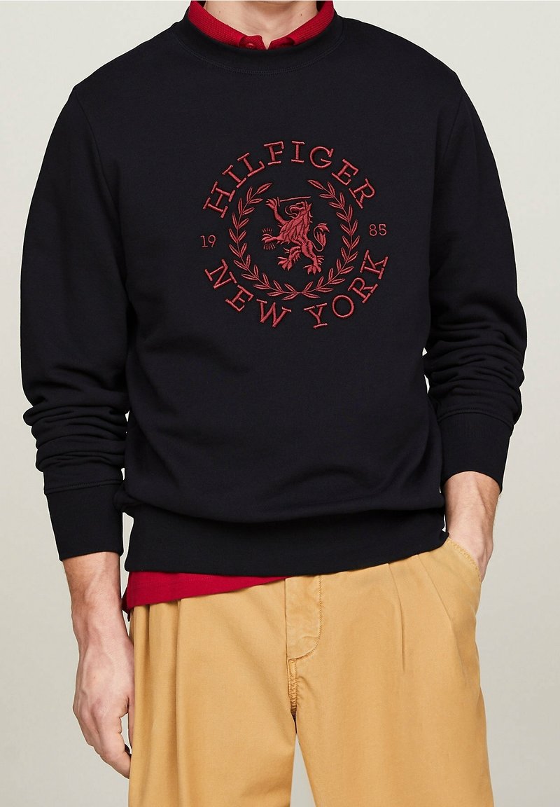 Homme portant un sweat-shirt noir avec un emblème rouge "Hilfiger New York 1985", une chemise rouge en dessous et un pantalon beige, une main dans la poche.