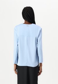 Blusa azzurra a maniche lunghe con scollo rotondo, dettaglio delle pences posteriori e un sottile logo vicino allo scollo. Tessuto con una texture liscia.