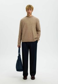 Hellbrauner Wollpullover mit geripptem Saum, kombiniert mit dunkelblauen, taillierten Hosen. Trägt eine dunkelblaue, weiche Tasche, braune Schuhe sichtbar.