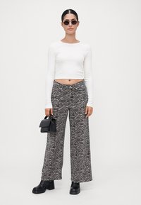 Femme portant un crop top blanc à manches longues, un pantalon large à motif zèbre, des bottes noires, des lunettes de soleil et tenant un petit sac à main noir sur un fond uni.