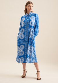 Abito lungo a maniche lunghe blu con motivi paisley bianchi, chiusura a bottoni frontale e tasche laterali. Tessuto morbido con una vestibilità comoda. Sandali neri con tacco.