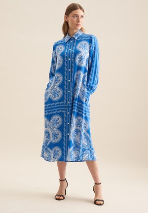 Abito lungo a maniche lunghe blu con motivi paisley bianchi, chiusura a bottoni frontale e tasche laterali. Tessuto morbido con una vestibilità comoda. Sandali neri con tacco.