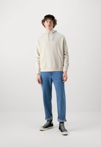 Krämfärgad hoodie med känguruficka, dragsko på huvan och ribbade ärmslut, kombinerad med ljusblå jeans och svarta högstövelsnickers.