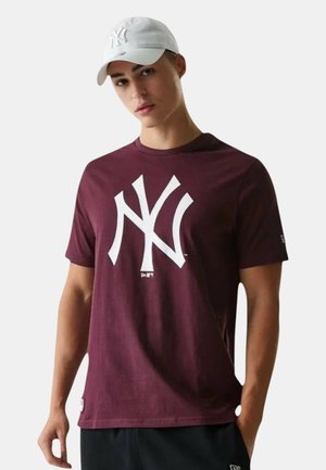 Joven con gorra blanca y camiseta burdeos con un gran logo blanco de los New York Yankees, de pie frente a un fondo liso.