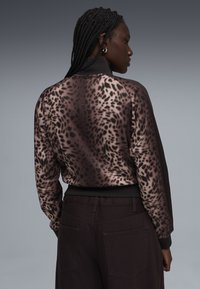 Γυναίκα με πλεγμένα μαλλιά που φοράει μπουφάν με λεοπάρ σχέδιο και σκούρο παντελόνι, στραμμένη πλευρικά σε γκρι φόντο με διαβαθμίσεις.