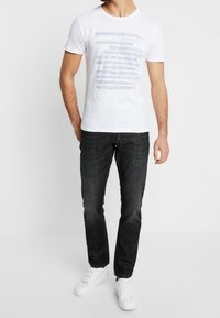 Hvid kortærmet t-shirt med blå grafiske striber, parret med mørke jeans og hvide sneakers; afslappet pasform og enkelt design.