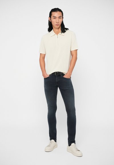 Φωτεινό μπεζ πόλο μπλουζάκι κοντού μανικιού με κλείσιμο φερμουάρ, συνδυασμένο με σκούρο μπλε skinny τζιν και λευκά αθλητικά παπούτσια. Μινιμαλιστικός σχεδιασμός, εφαρμοστό στυλ.