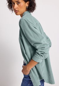 Donna con capelli ricci corti, che indossa orecchini a cerchio, una maglietta a coste verde chiaro e jeans blu, rivolta di lato con le mani in tasca.