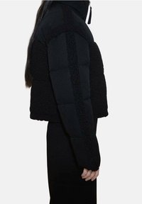 Kobieta w czarnej, krótkiej kurtce puchowej z teksturowanymi panelami, stojąca bokiem na tle jednolitej białej ściany.